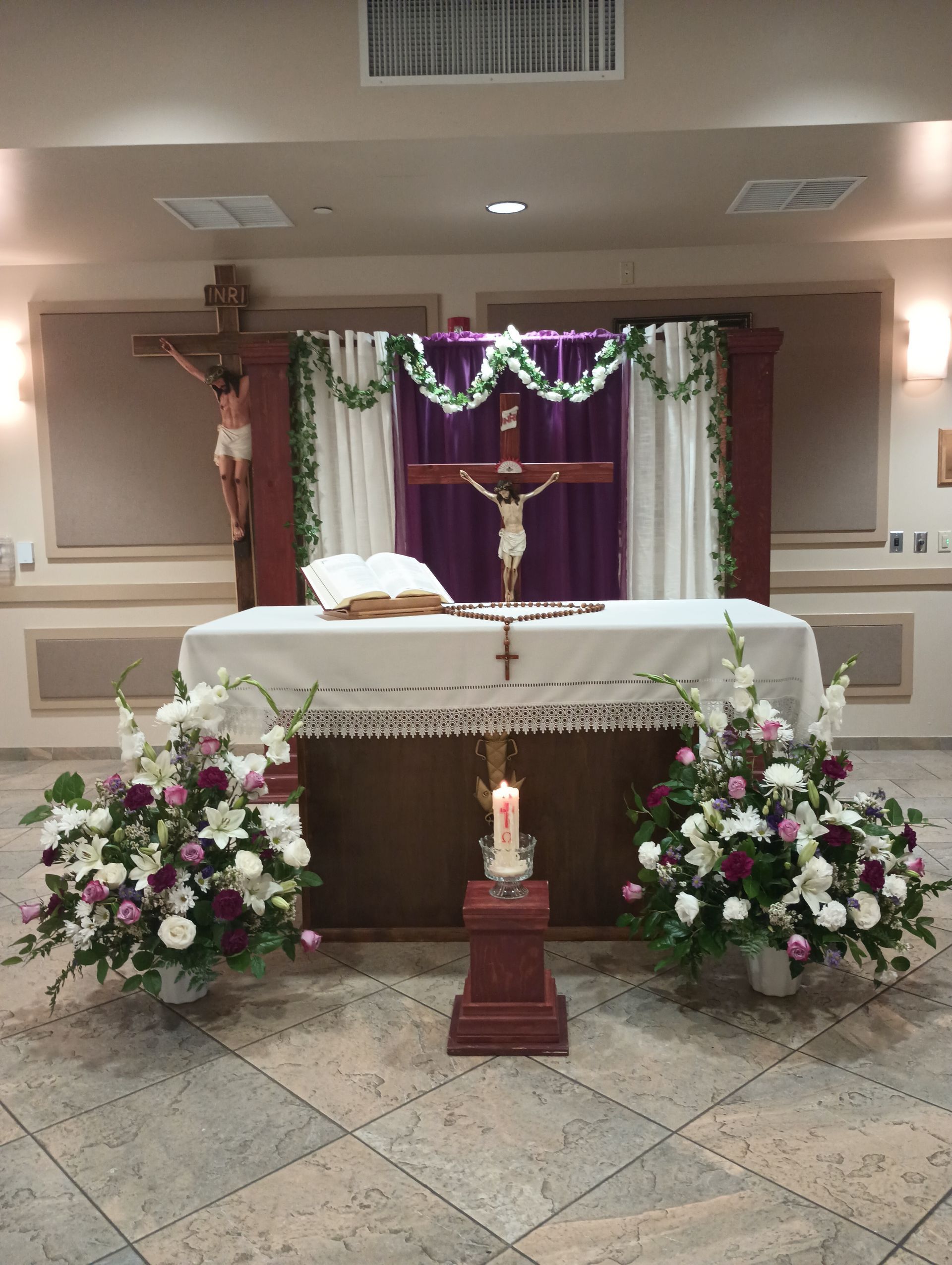 Church altar decorated with white cloth, purple drapery, candles, flowers, and a crucifix