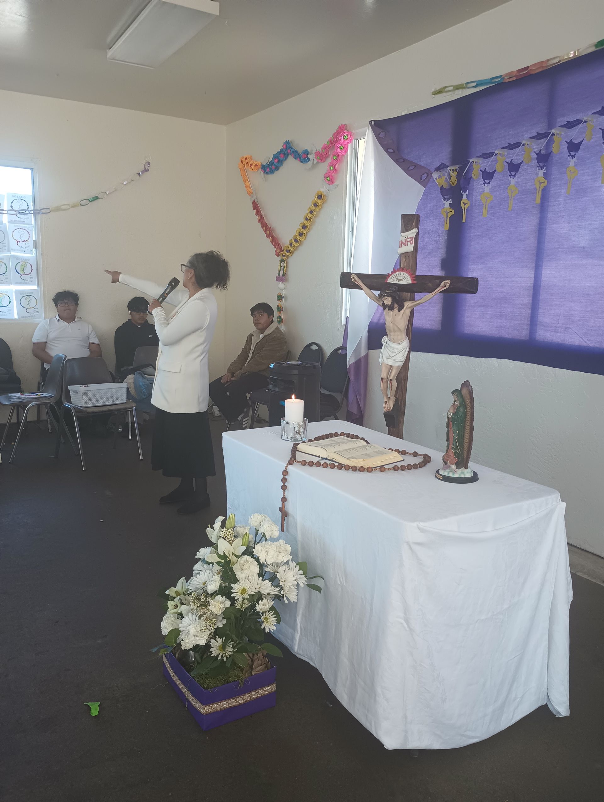 Small church altar with a crucifix, flowers, candles, and a person speaking near seated attendees.