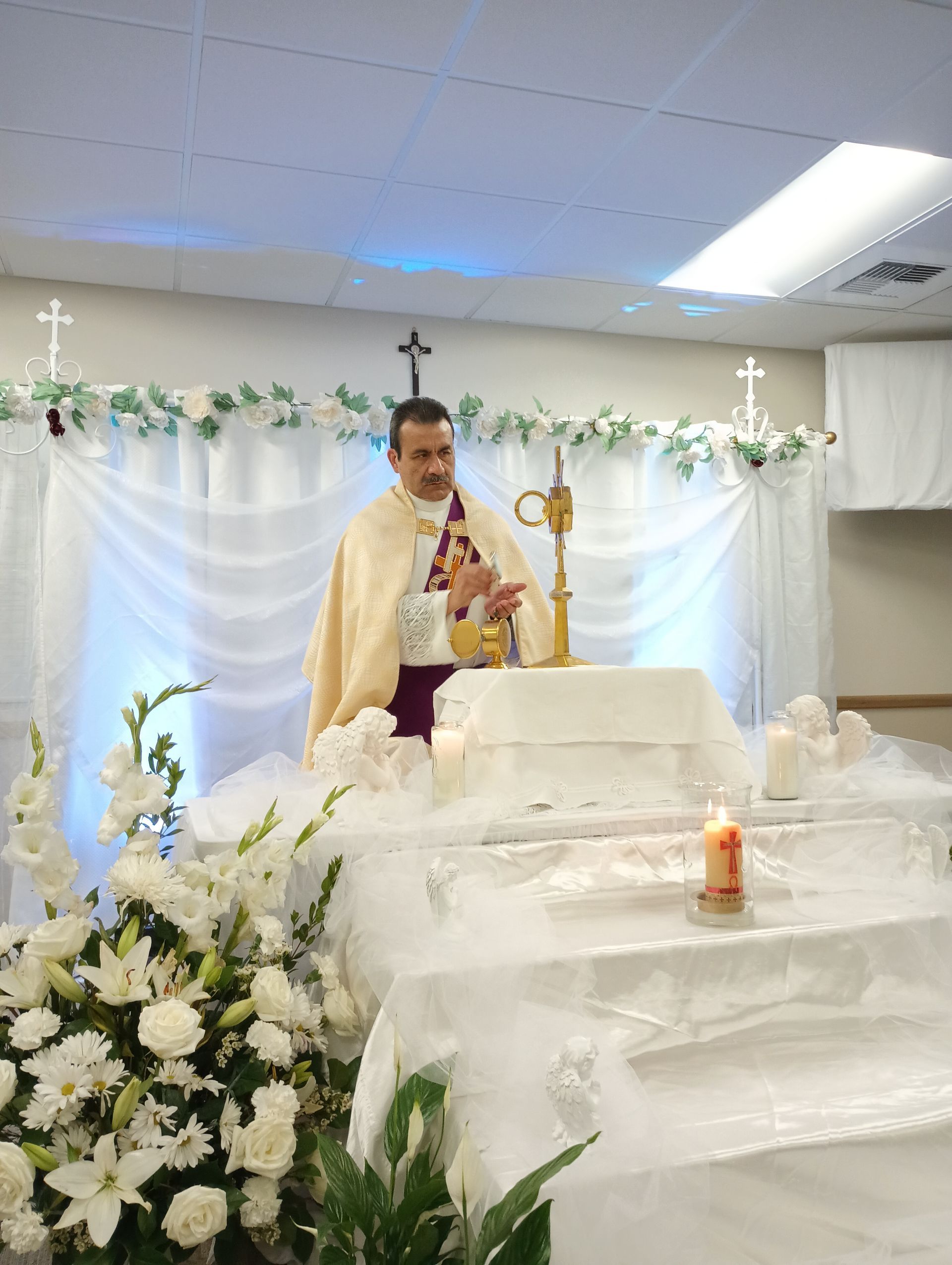Clergyman at an altar decorated with white flowers and candles, holding a microphone during a church service
