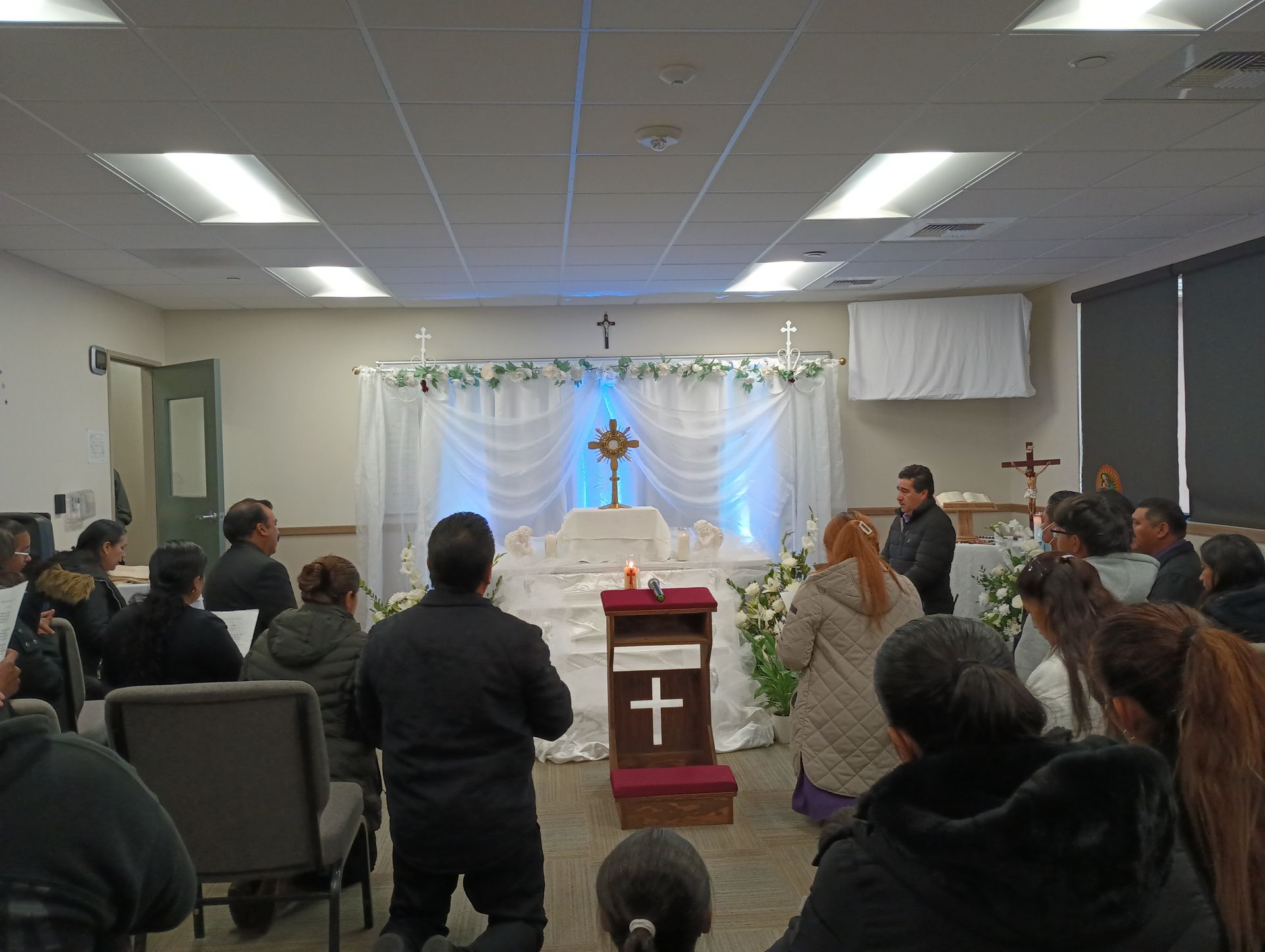 People seated in a small church service facing a decorated altar with a cross and white drapery.