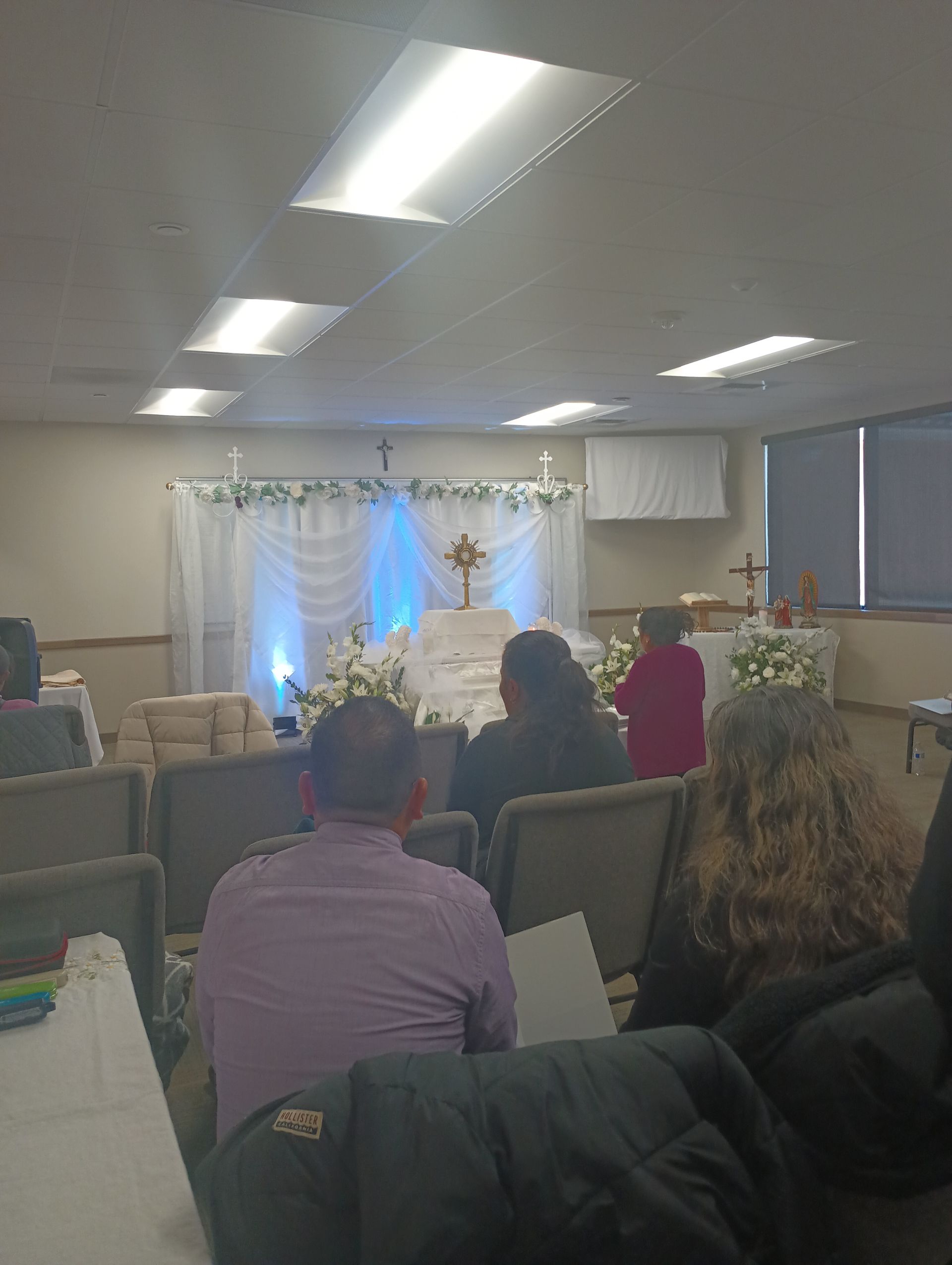 People seated in a small room watching a decorated stage with white drapes and blue lighting.