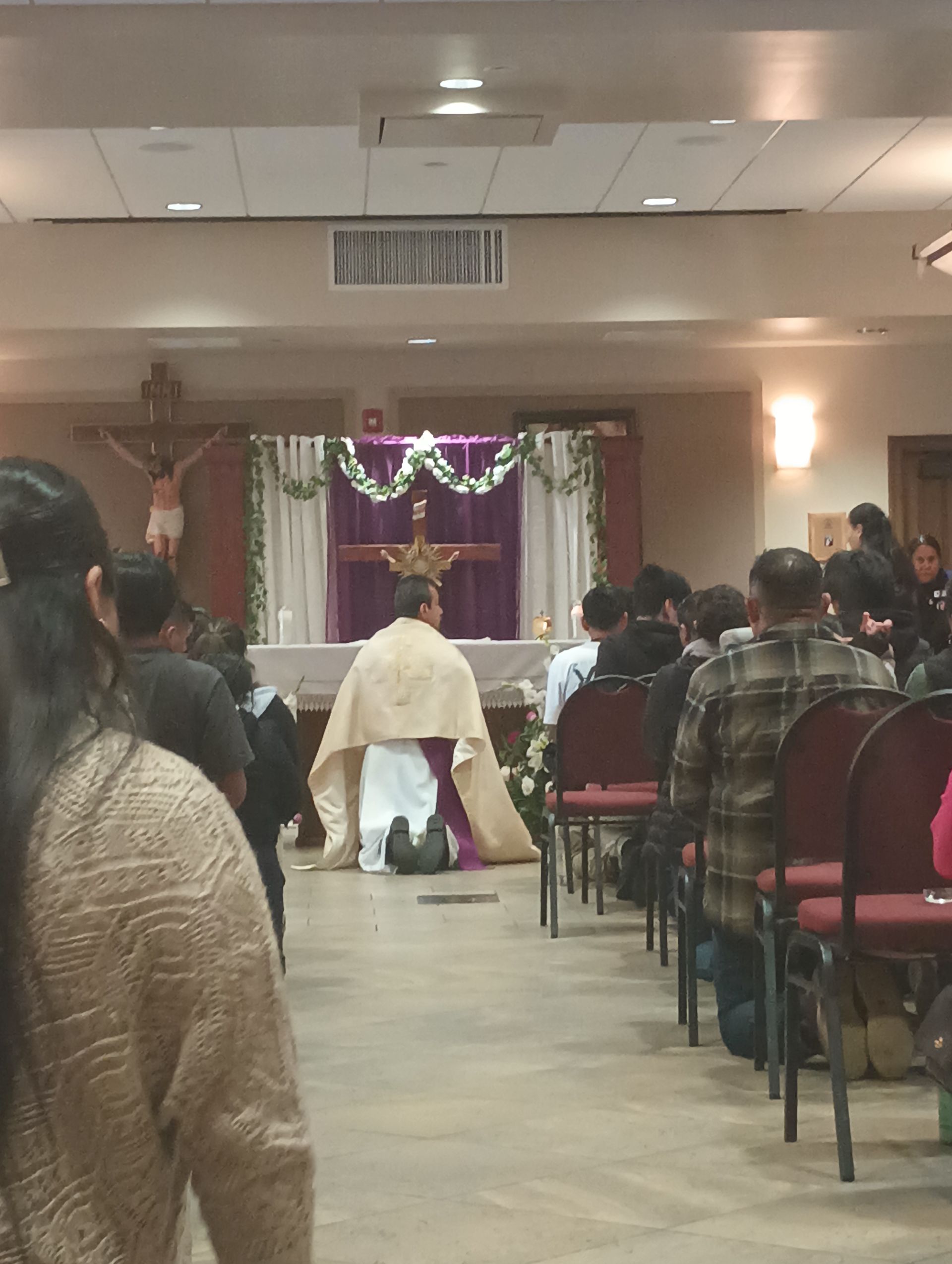 Indoor church ceremony with people seated facing a decorated altar and a person standing at the front