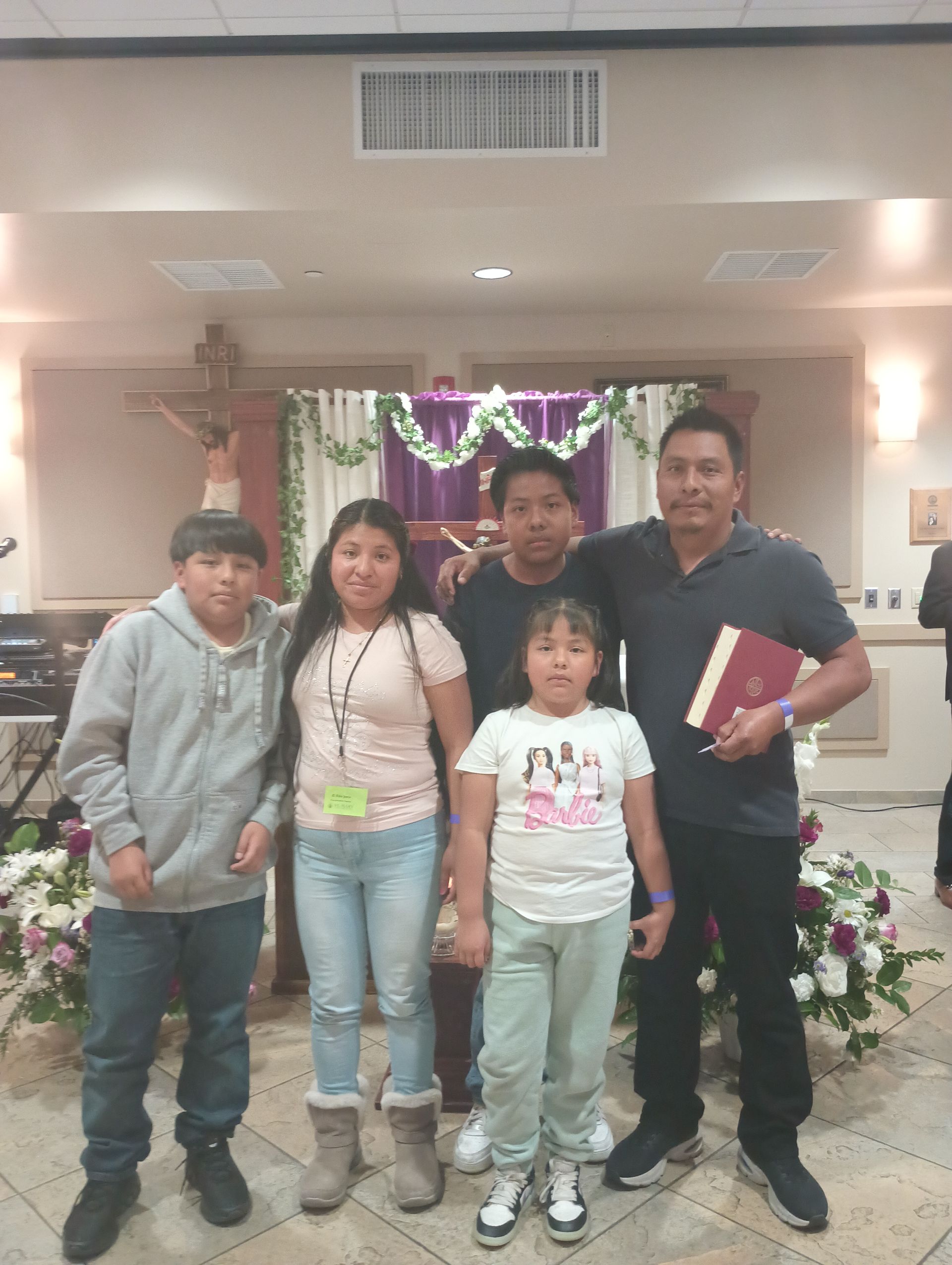 Five people posing indoors at a decorated event, with flowers and a stage in the background.
