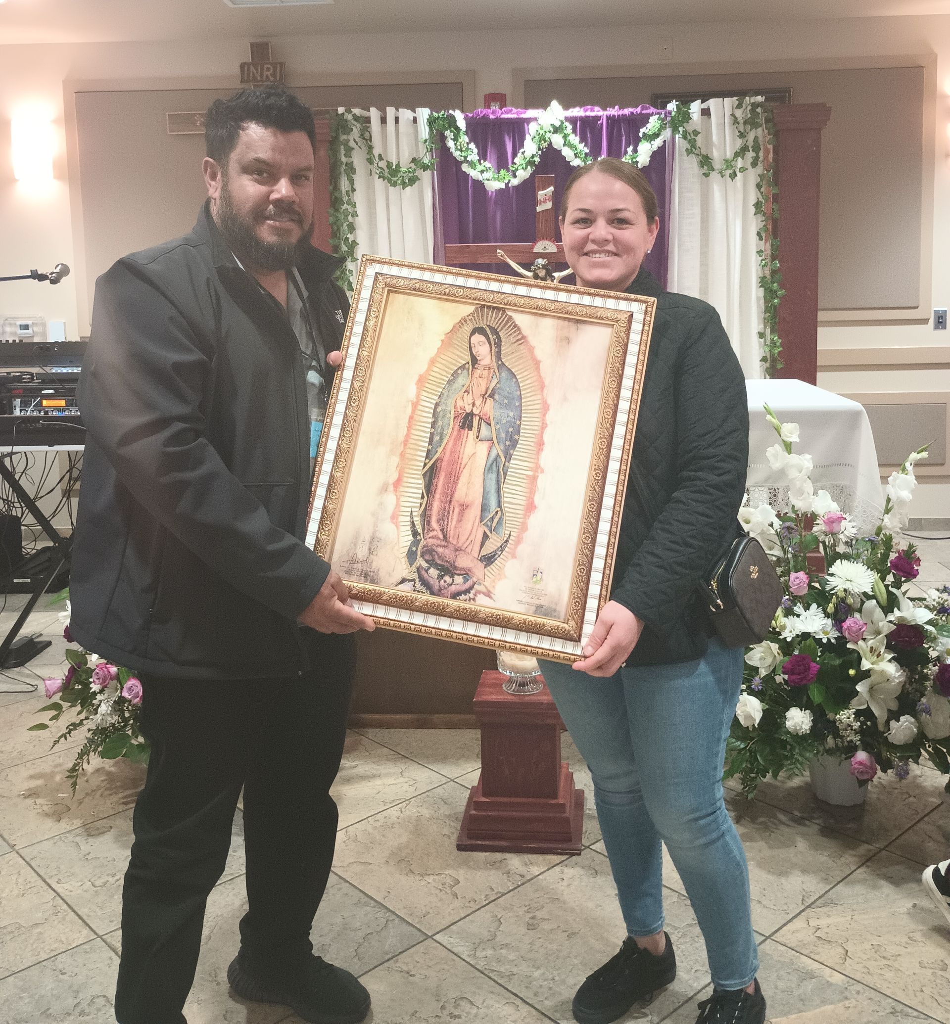 Two people holding a framed image of the Virgin Mary in a decorated indoor hall