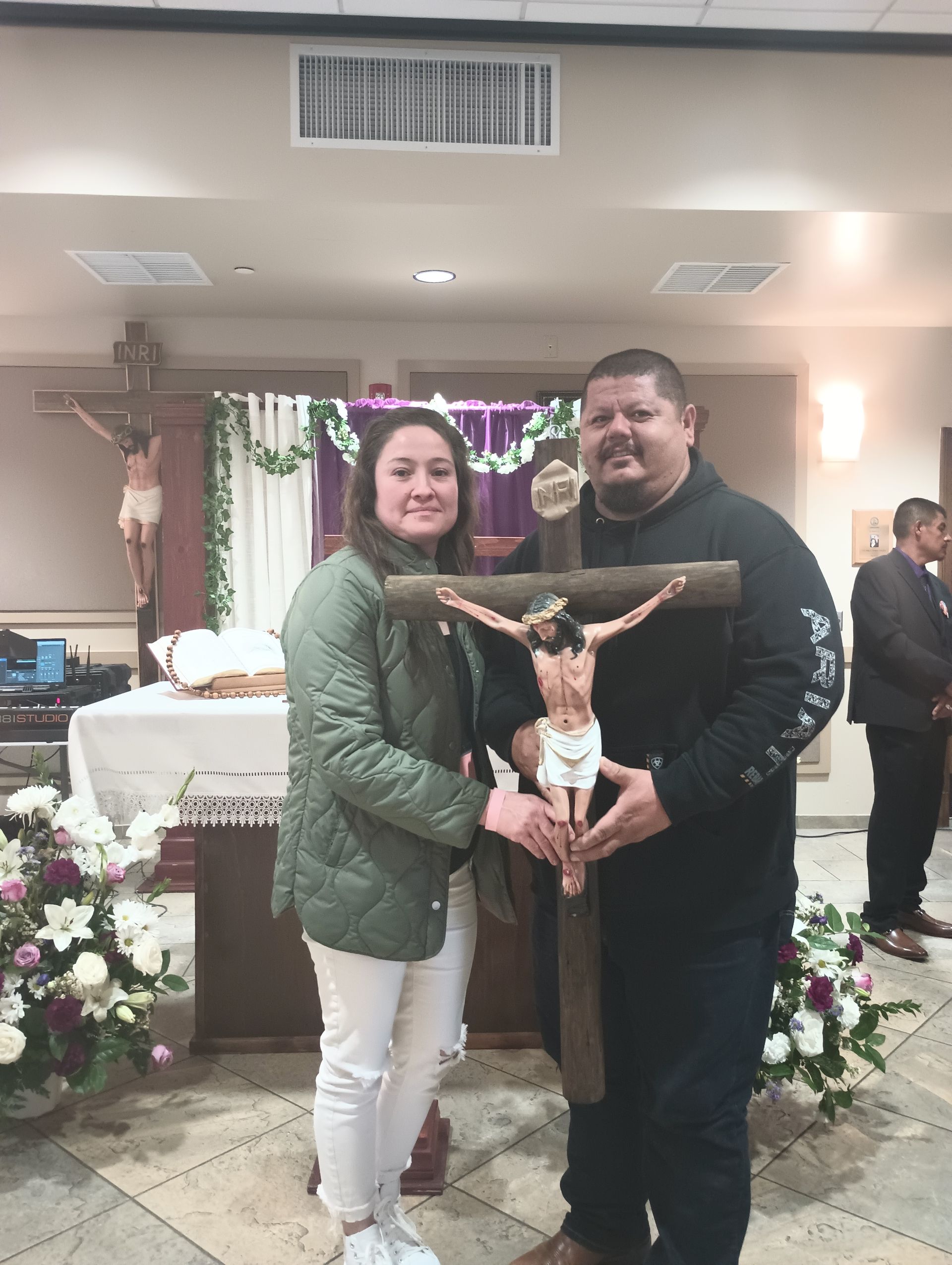 Couple posing beside a crucifix in a decorated church hall, with floral arrangements behind them.