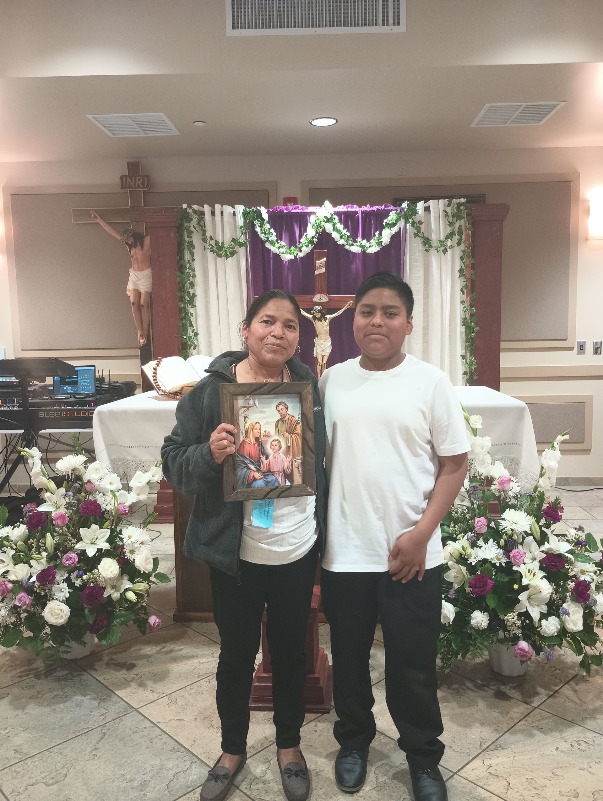 Two people pose in a decorated church, holding a framed photo before an altar with flowers and purple drapery.