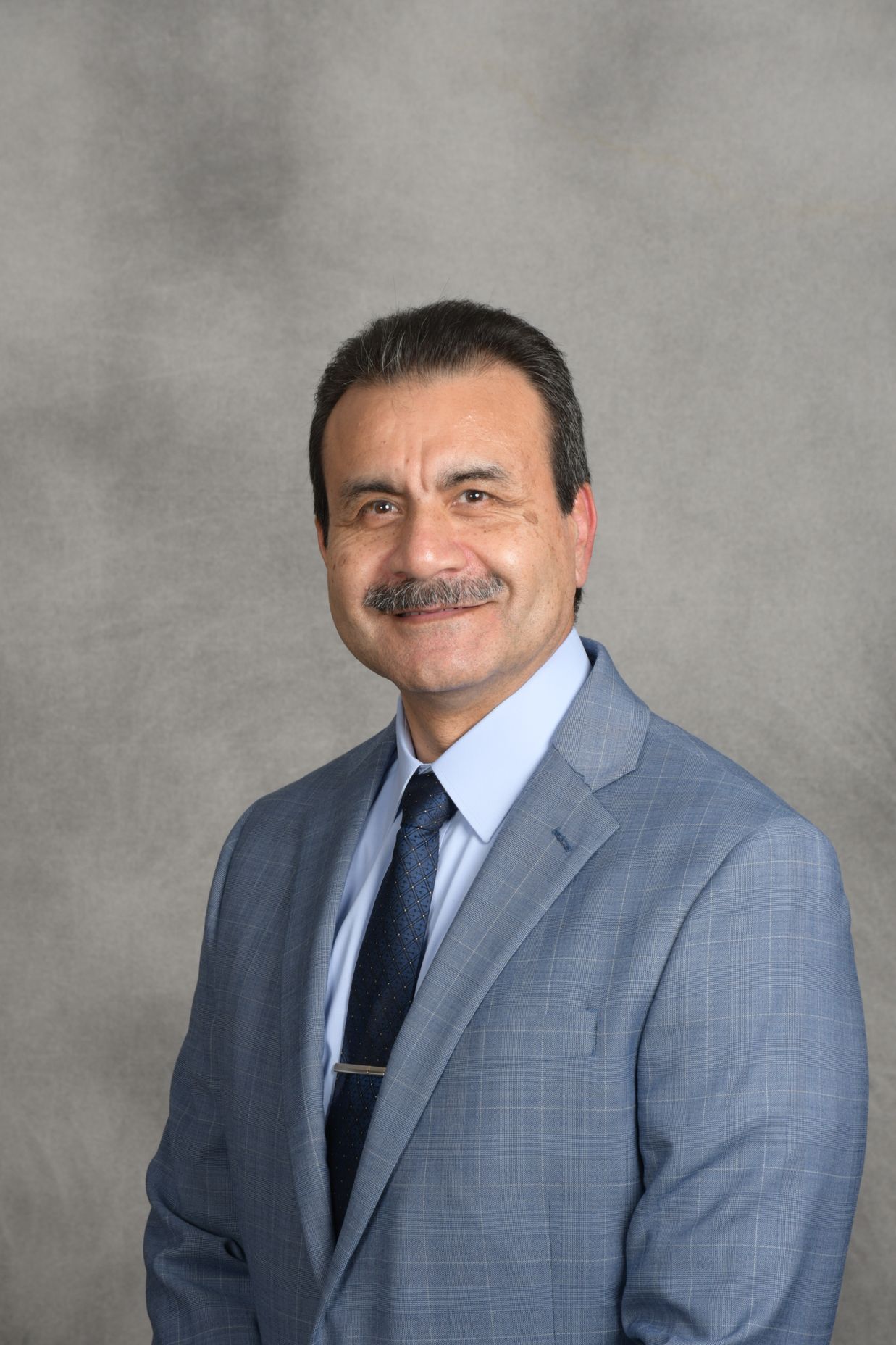 Portrait of a man in a light blue suit and tie, standing against a gray studio backdrop.
