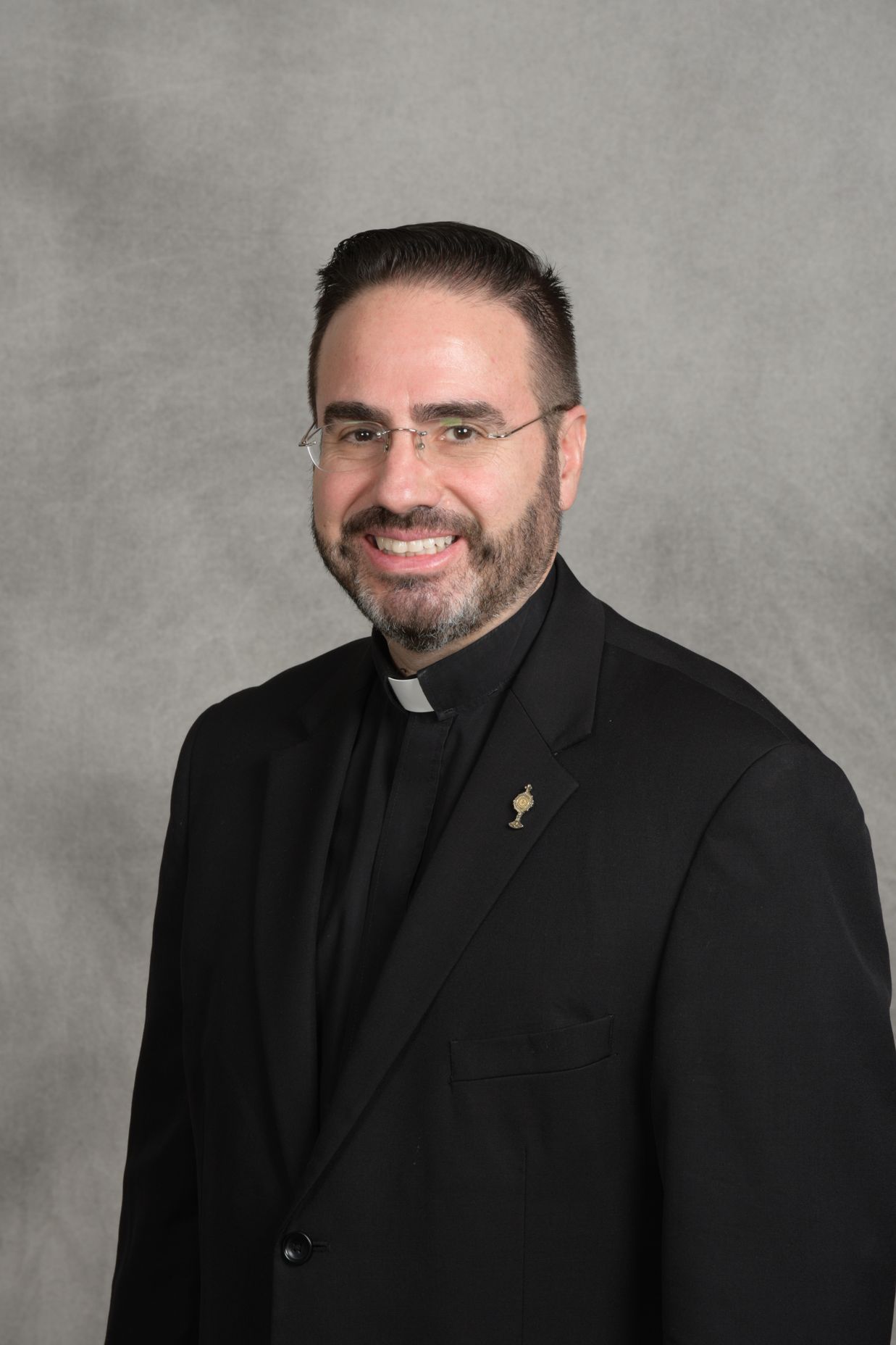 Smiling man in black clerical attire and glasses against a gray studio backdrop