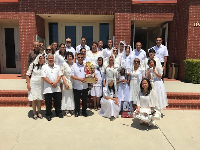 Group of people in white outfits posing outside a brick building, holding bouquets and a gift bag.