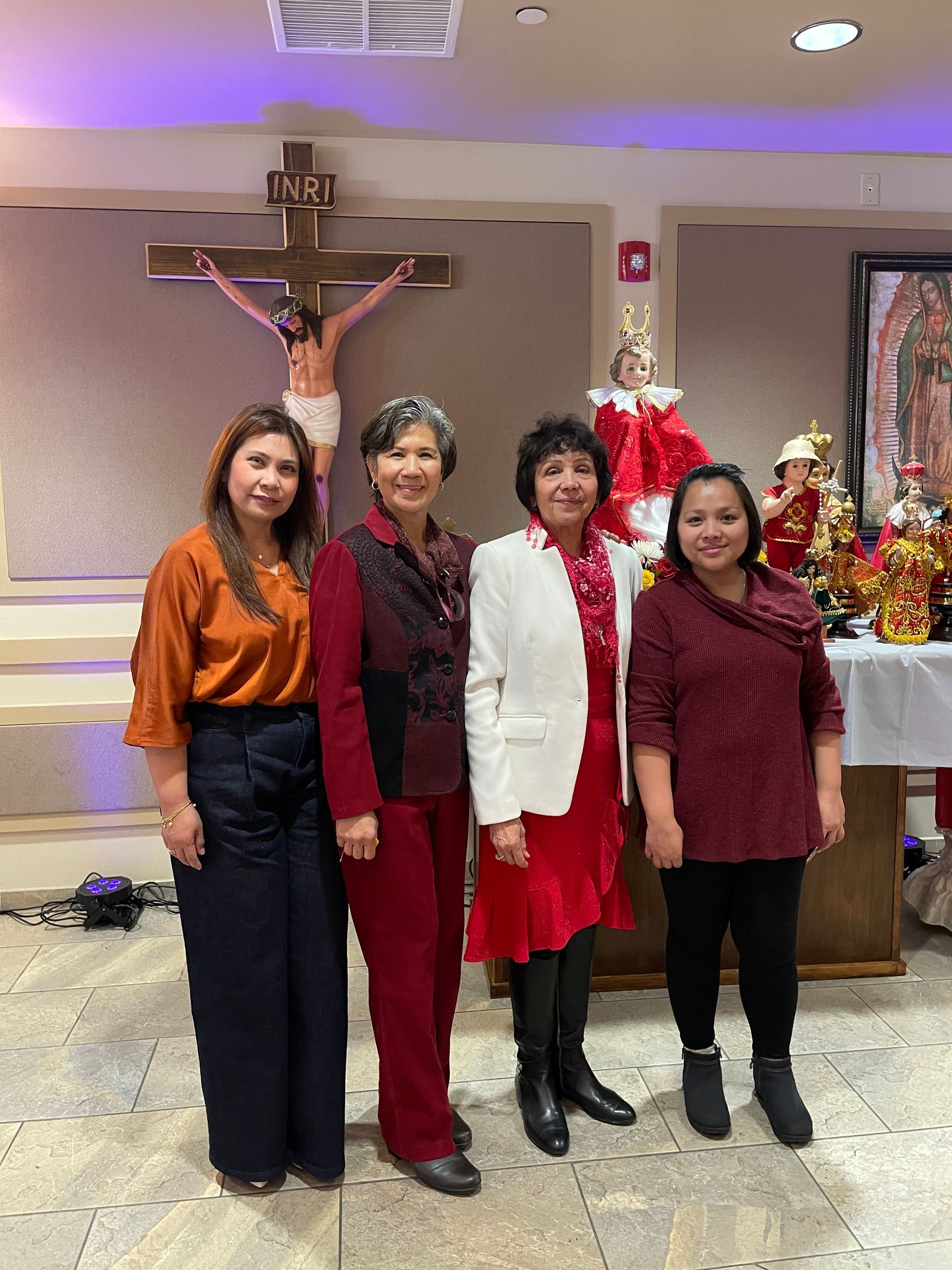 Four people stand indoors before a crucifix, dressed in festive red, white, and black outfits.