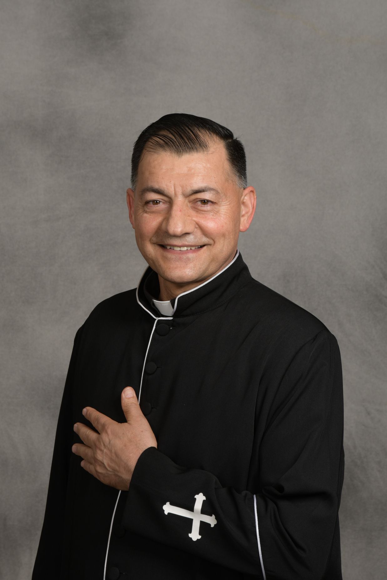 Portrait of a smiling man in a black clerical shirt with a white collar, against a gray background
