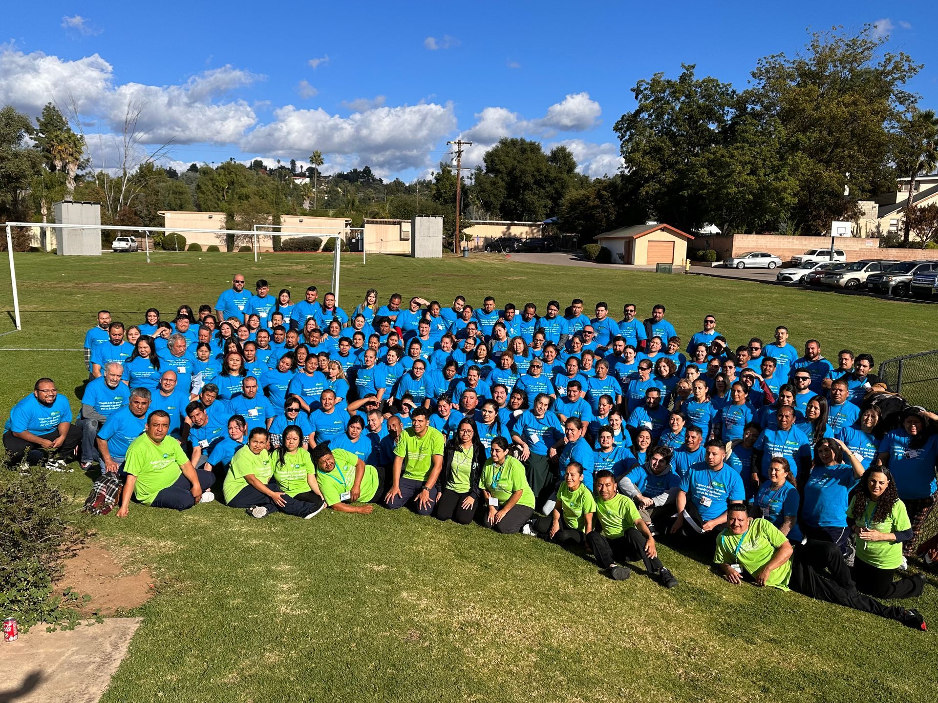 Large group posing outdoors in blue and neon green shirts on a grassy field under a bright sky