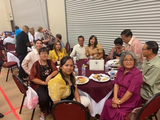 Group seated at banquet tables in a hall, chatting around maroon tablecloths and plates of food.