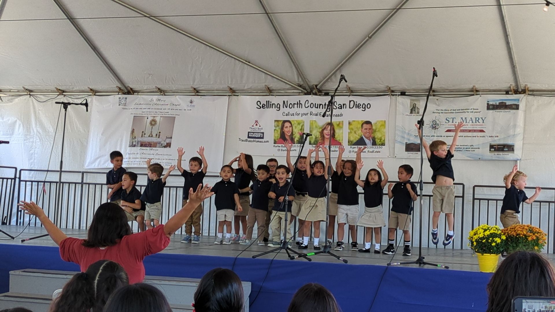 Children performing on a stage under a white tent, with raised arms and banners behind them.