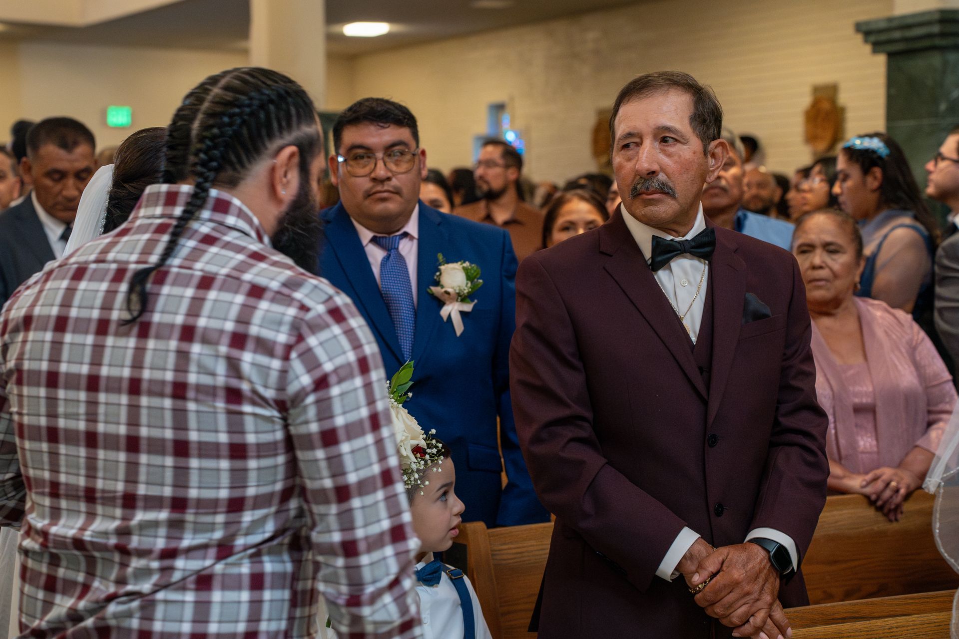 People seated at an indoor ceremony; a man in a burgundy suit stands in the foreground.