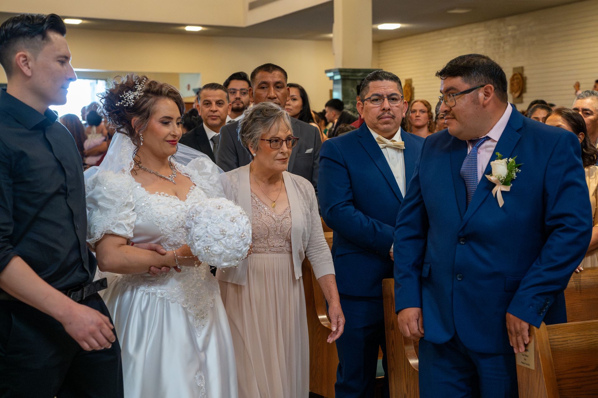 Wedding party gathered indoors; bride in white stands beside guests in blue suits and an older woman.
