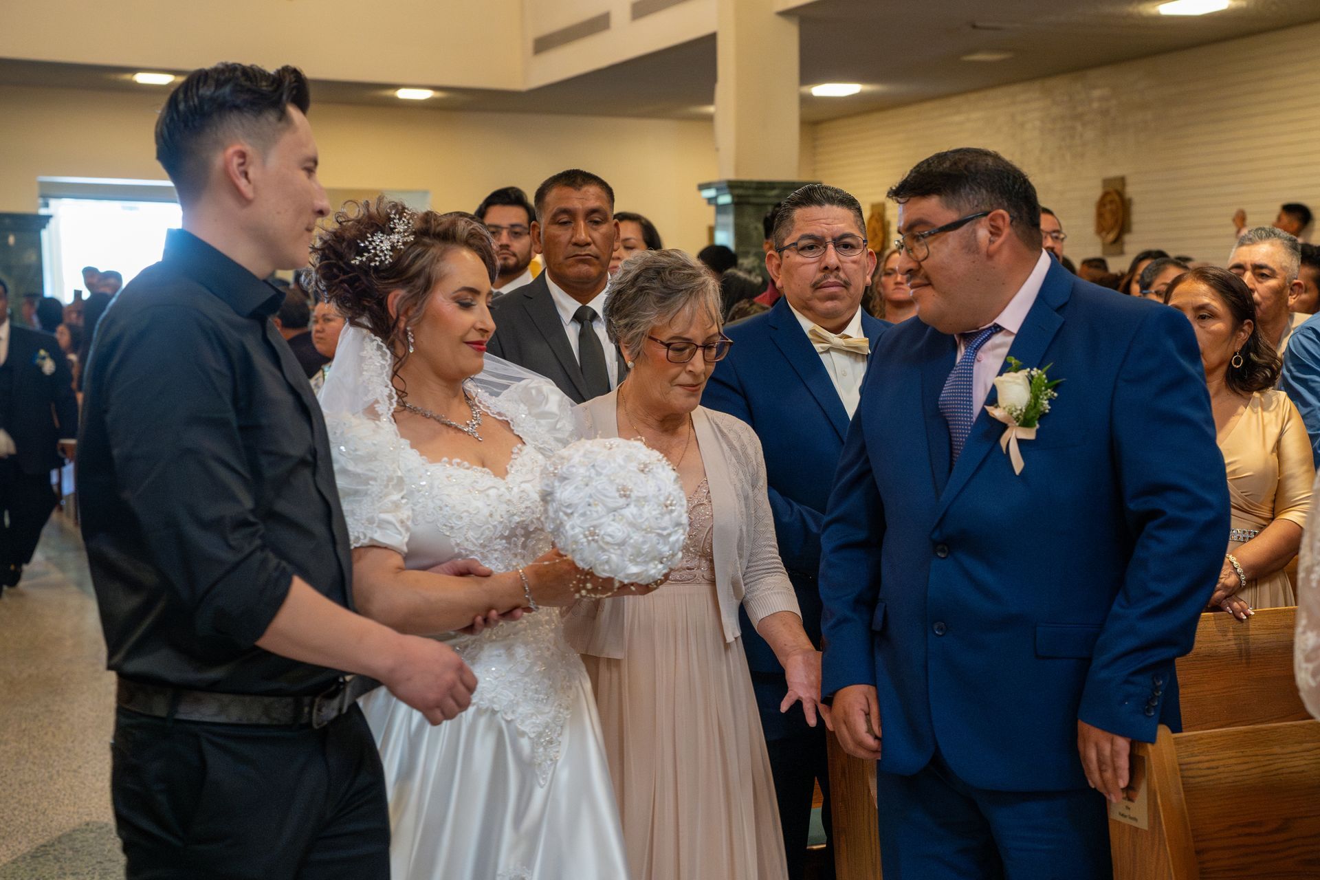 Wedding party gathered indoors, with bride in white holding bouquet and groom in blue suit facing each other.