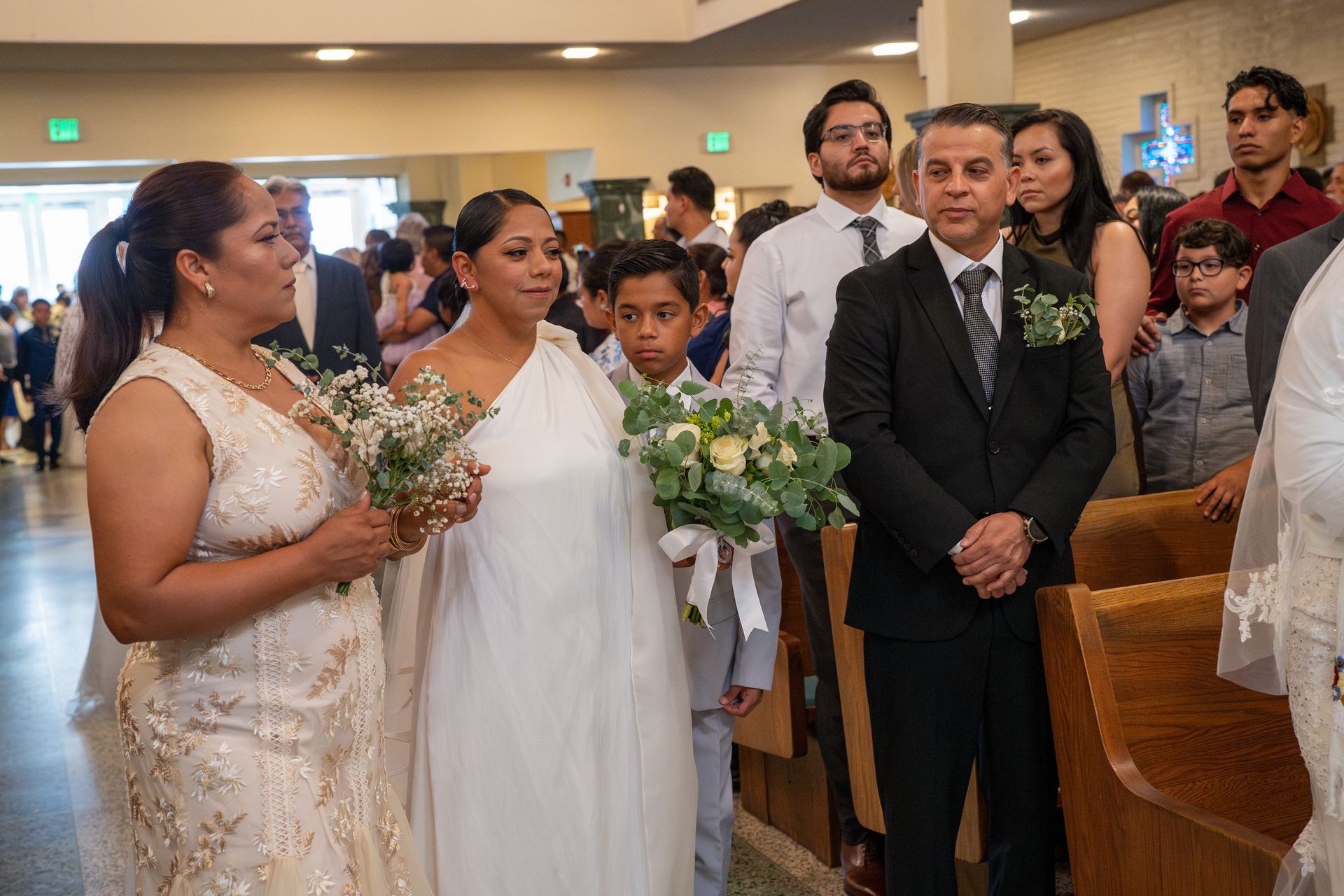 Wedding guests and attendants standing in a church aisle, holding bouquets during the ceremony.