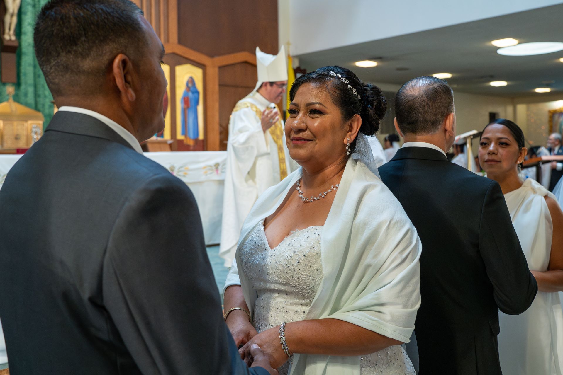 Bride in white dress and shawl at a church ceremony, facing a man in a suit with guests and clergy behind them