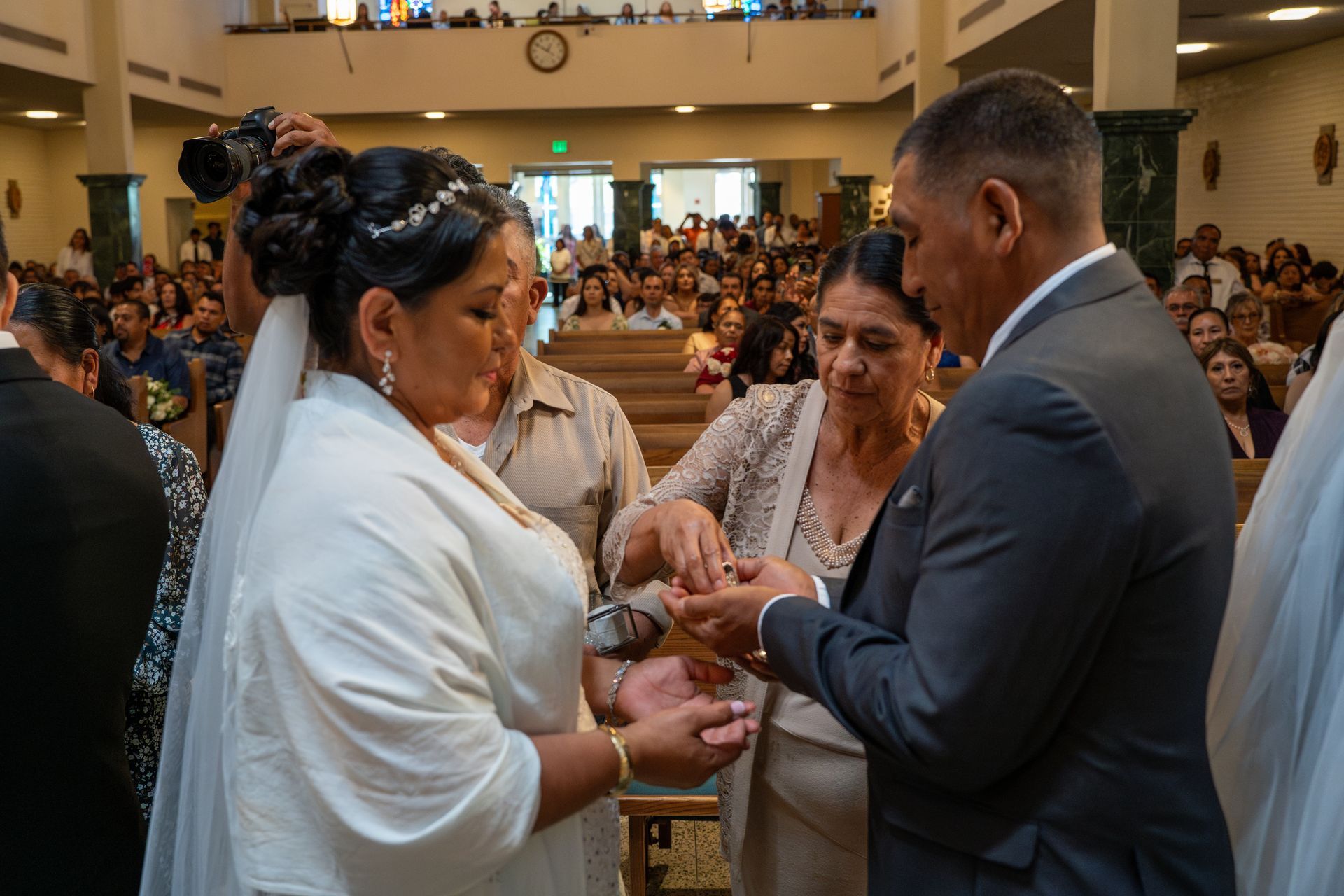 Couple exchanging rings during a church ceremony, with a crowded congregation in the background.