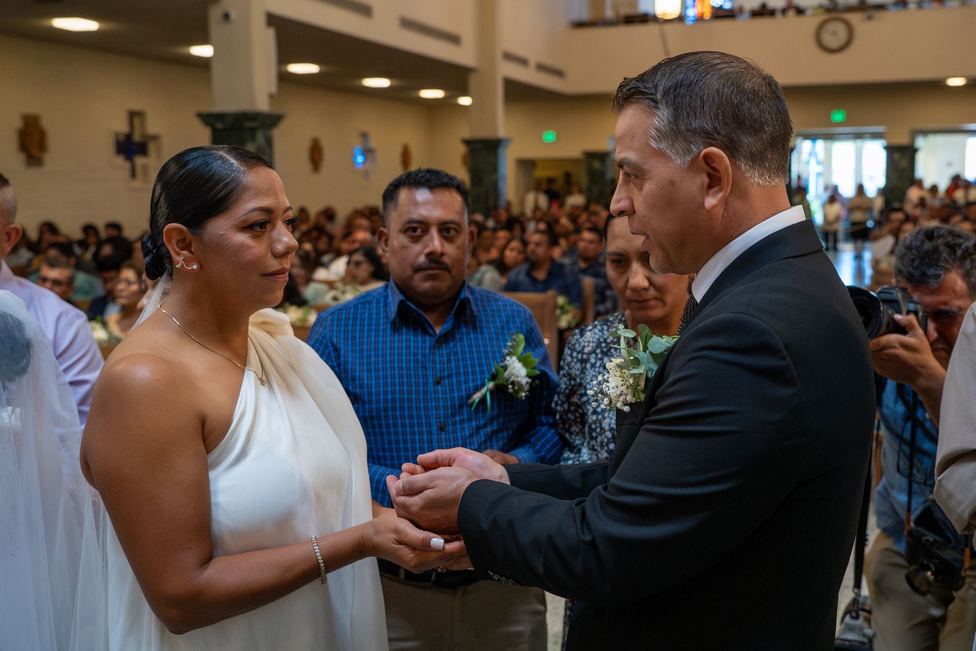 Two people in formal wear hold hands during a ceremony, with a crowd watching in the background.