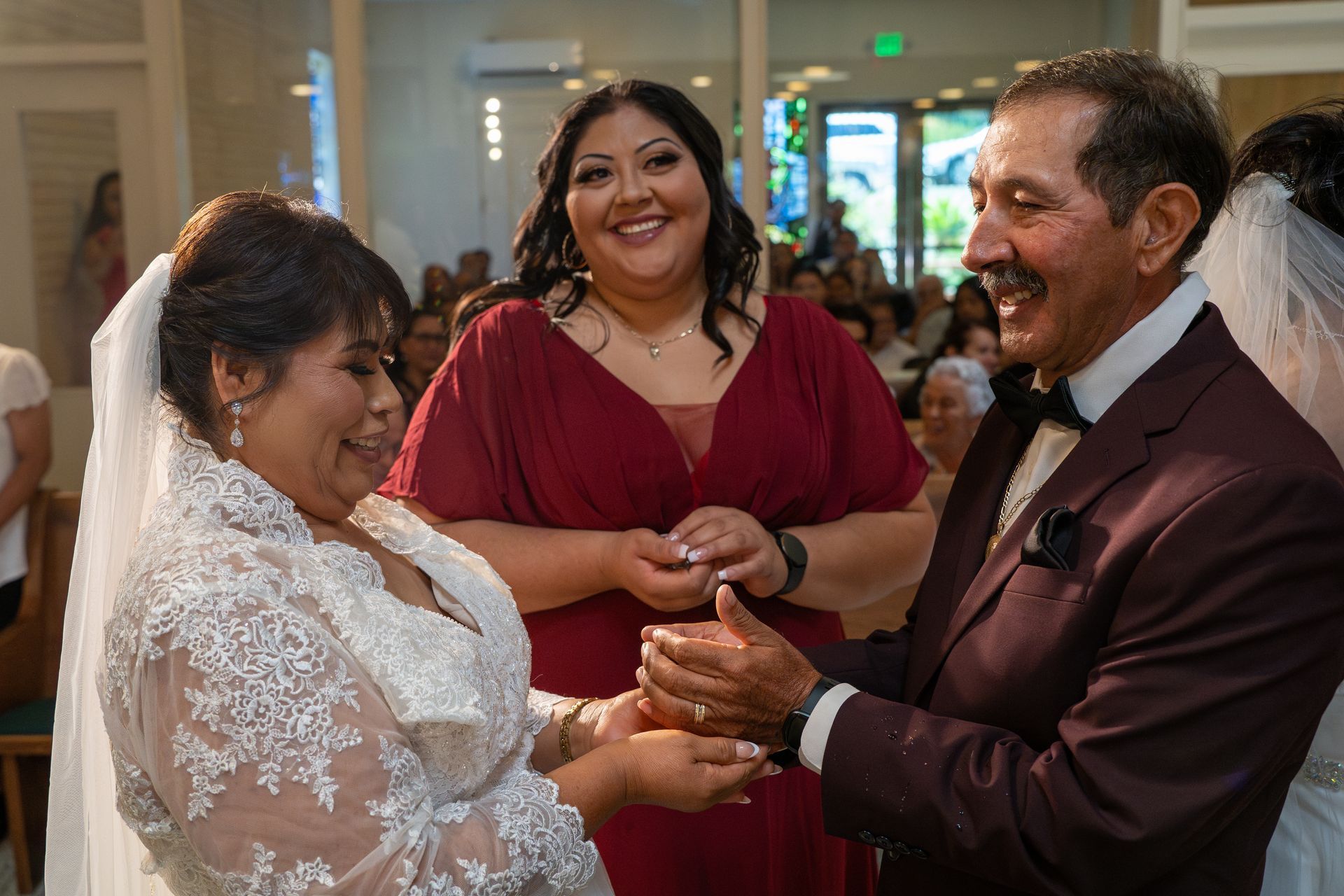 Bride and groom exchanging rings, with a smiling guest at an indoor wedding ceremony.