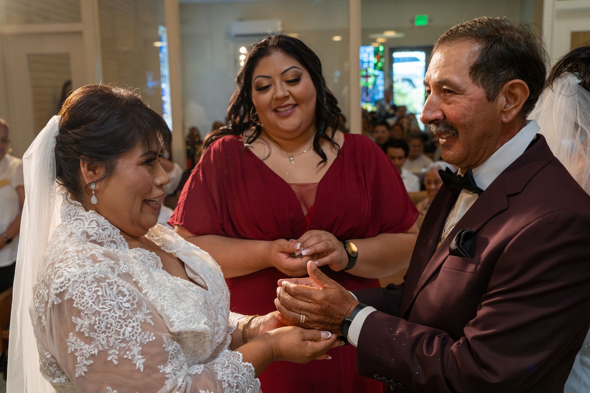 Bride and groom exchanging rings at a wedding ceremony, with a smiling guest in a red dress nearby