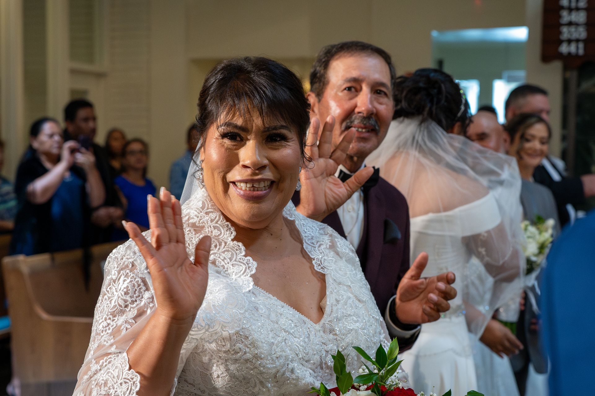 Woman in a white lace dress waving at a wedding ceremony, with guests smiling in the background