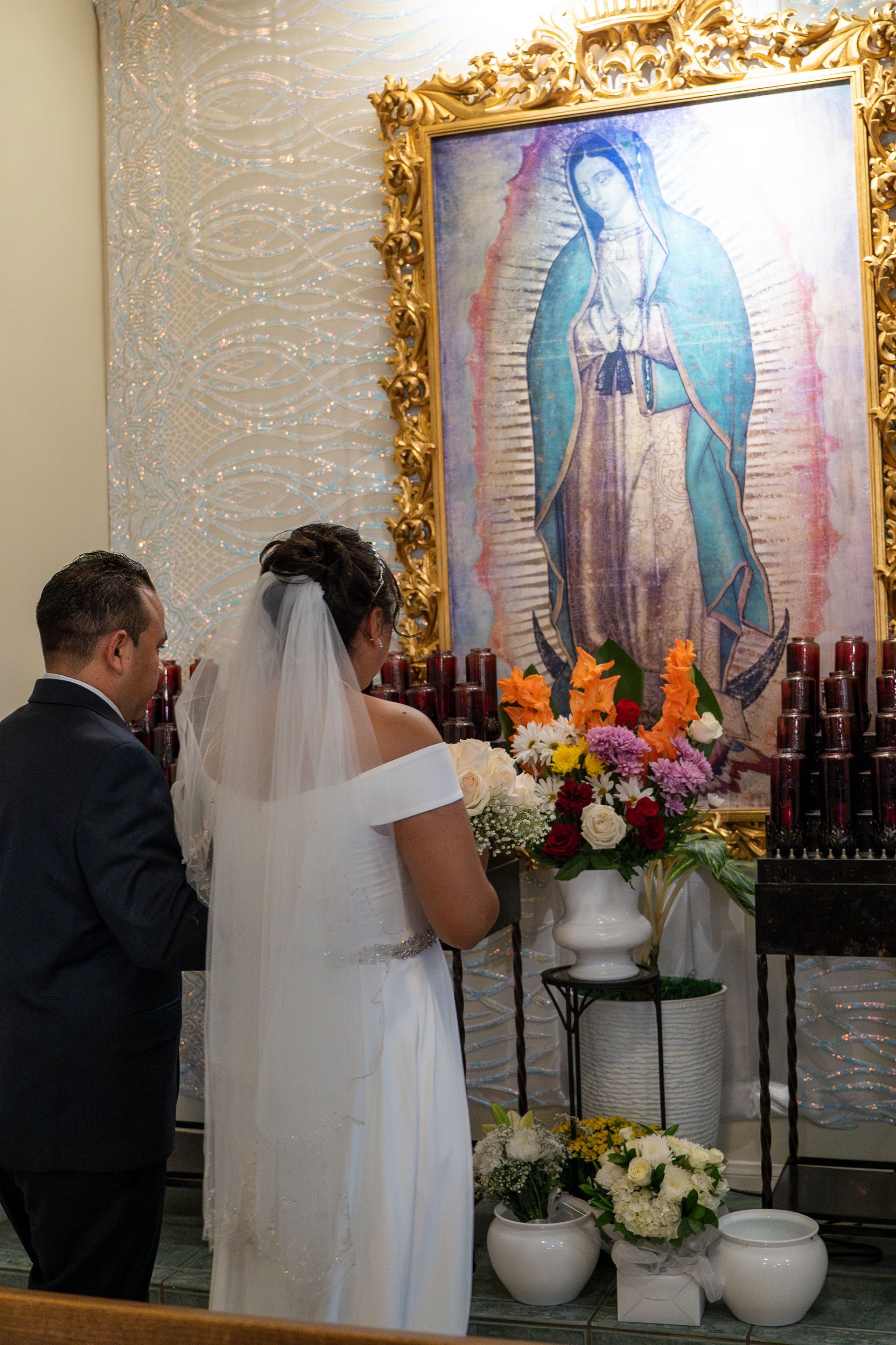 Couple standing before a framed Our Lady of Guadalupe altar surrounded by flowers and candles