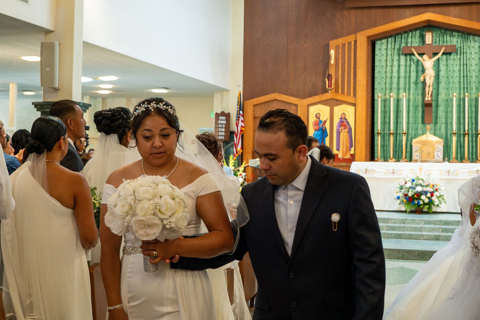 Wedding couple walking down church aisle, bride holding white bouquet beside groom in dark suit.