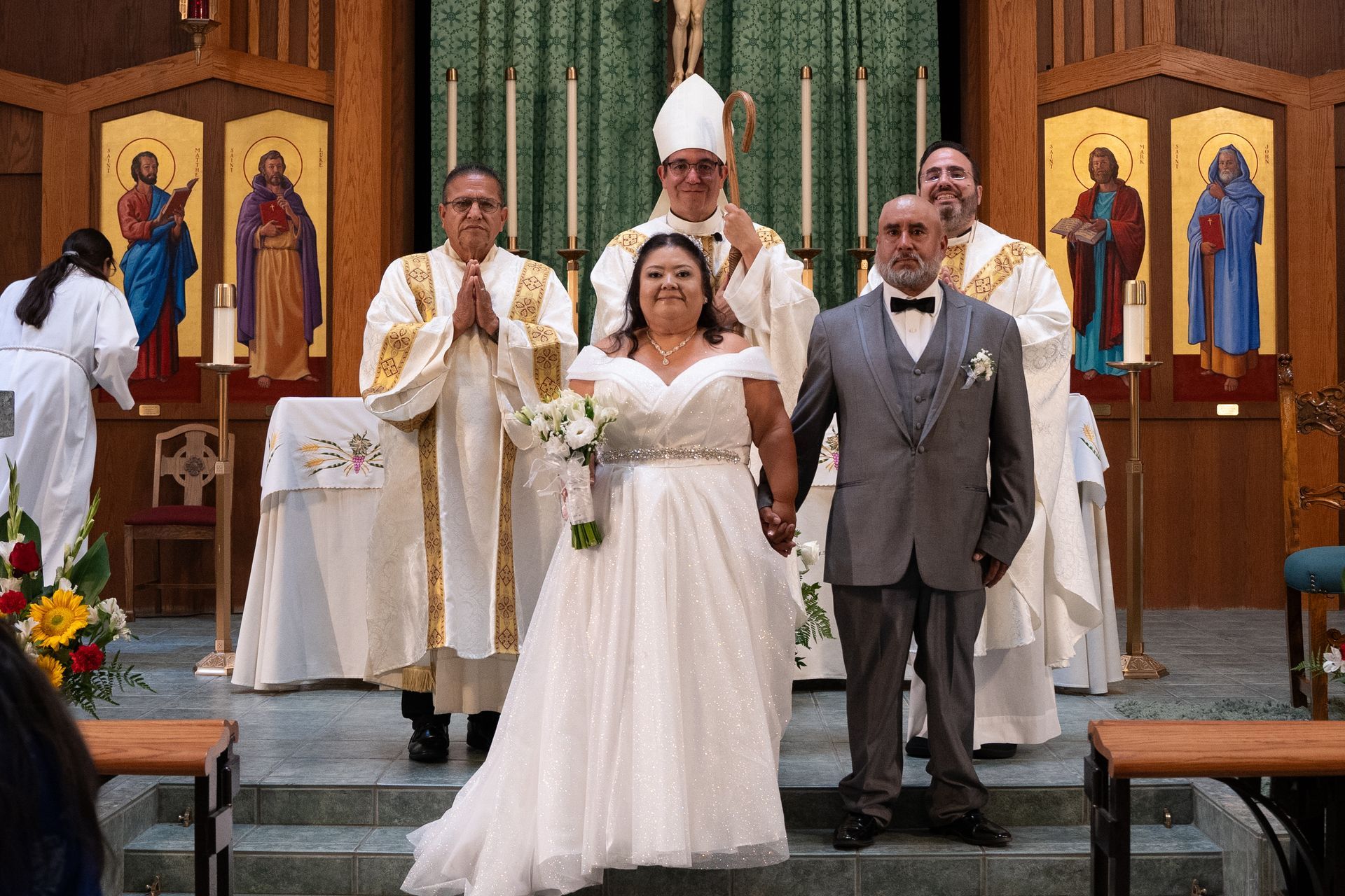 Wedding party posing at the altar in a church, with bride in white and groom in gray centered in front of a priest.