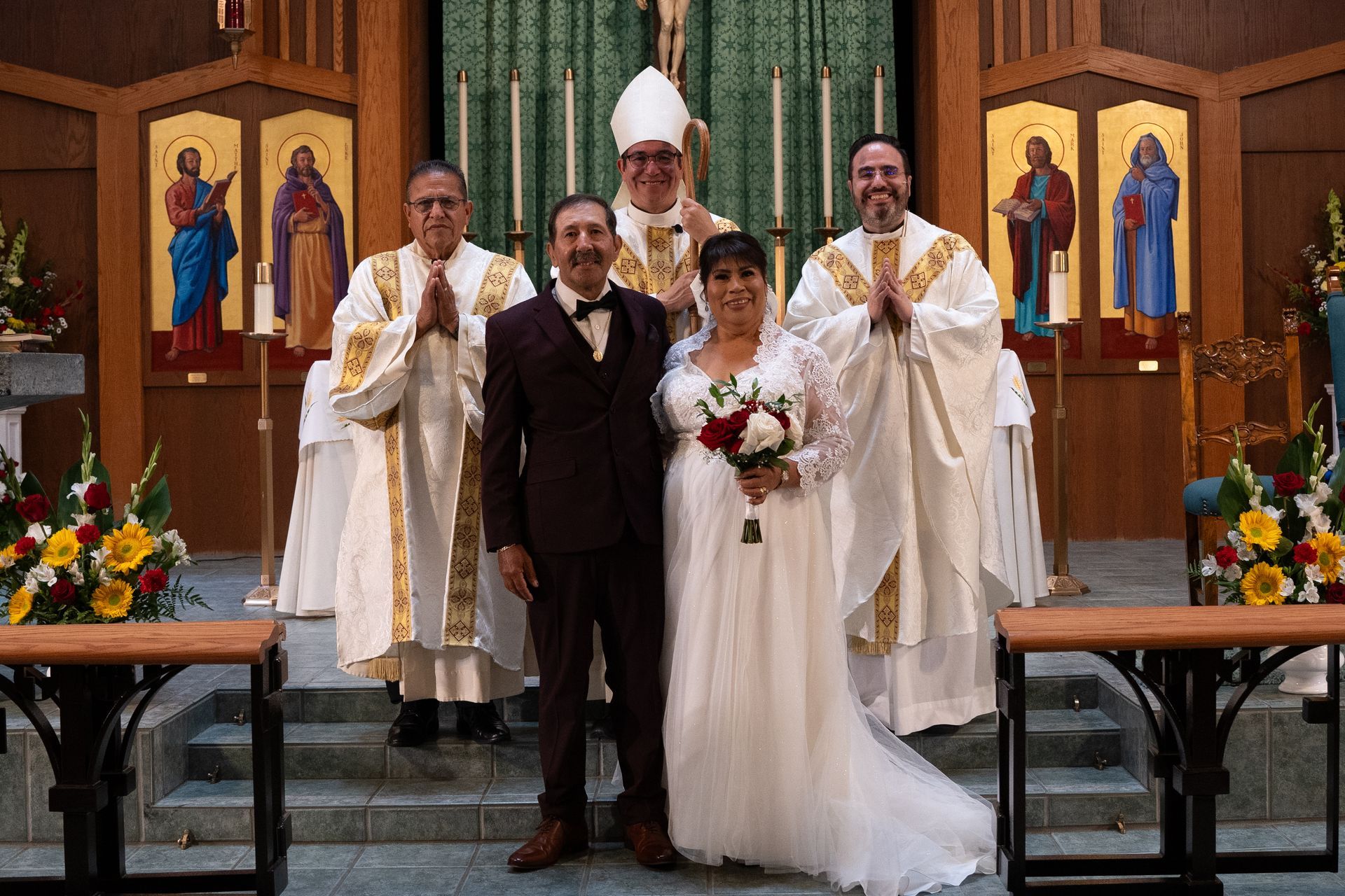 Wedding couple with priests in a church, standing at the altar with floral arrangements and stained glass.