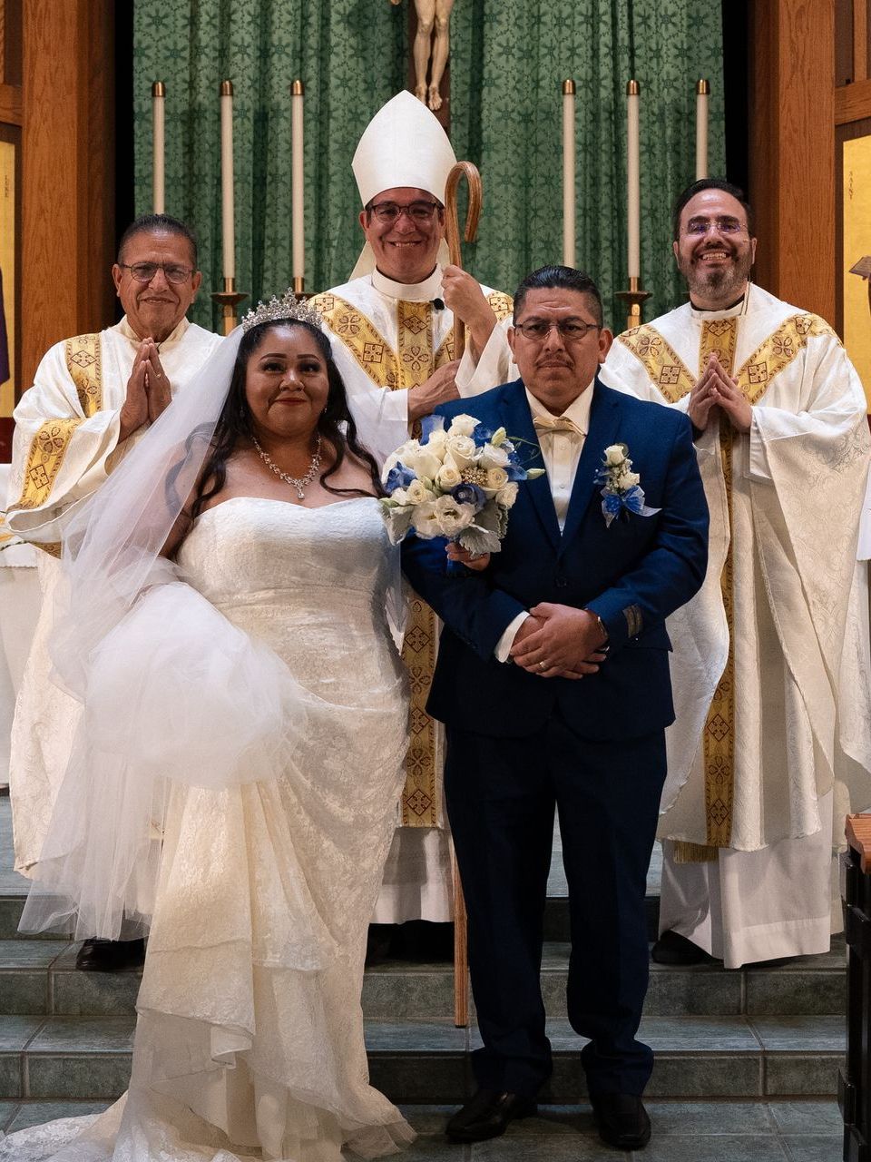 Bride and groom pose with clergy in a church, the bride in a white gown and the groom in a navy suit.