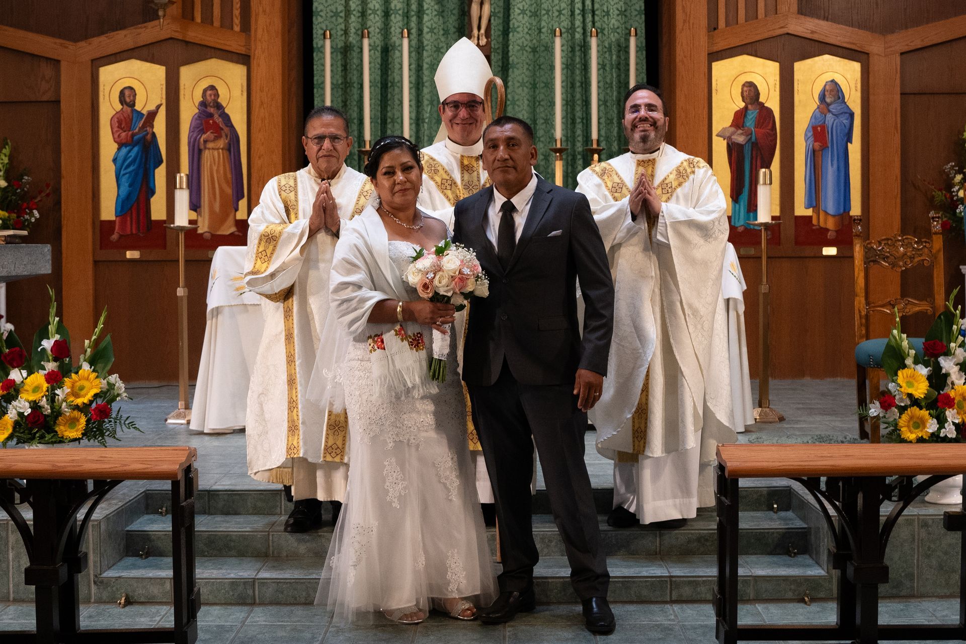 Wedding couple posing in a church with clergy behind them, holding a bouquet and bouquet of white flowers.