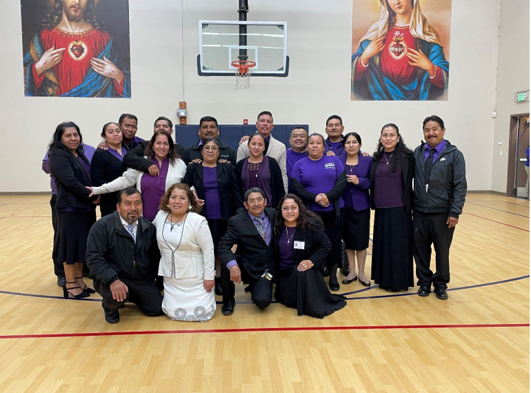 Group of people posing in a gymnasium under a basketball hoop, with colorful religious murals on the wall.