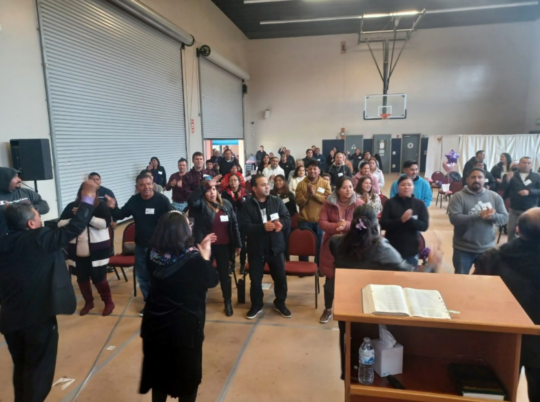 People gathered in a gym-like room, some clapping near a podium with an open book.