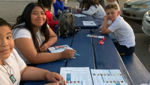 Students seated at a blue outdoor picnic table, smiling and working with papers and small objects