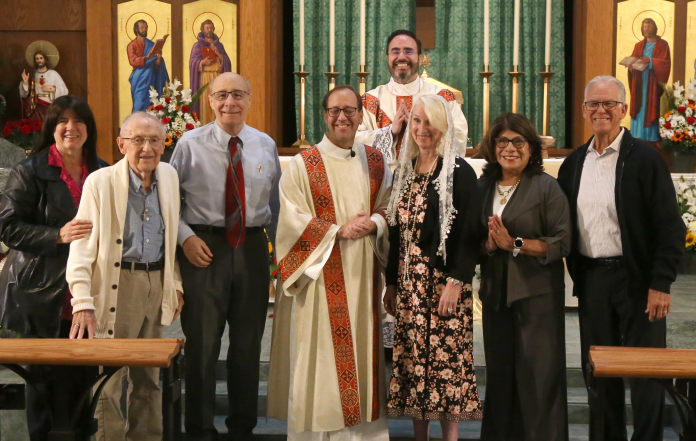 Seven people stand together in a church, smiling in front of stained glass windows.