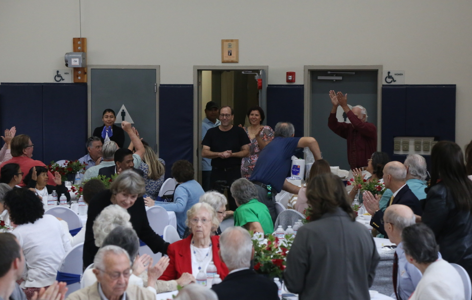 People gathered in a hall, sitting at tables and standing near doors, some clapping and raising hands.