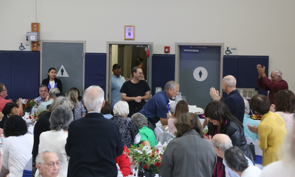 Crowded community hall with people seated and standing near a stage area and doors at the back