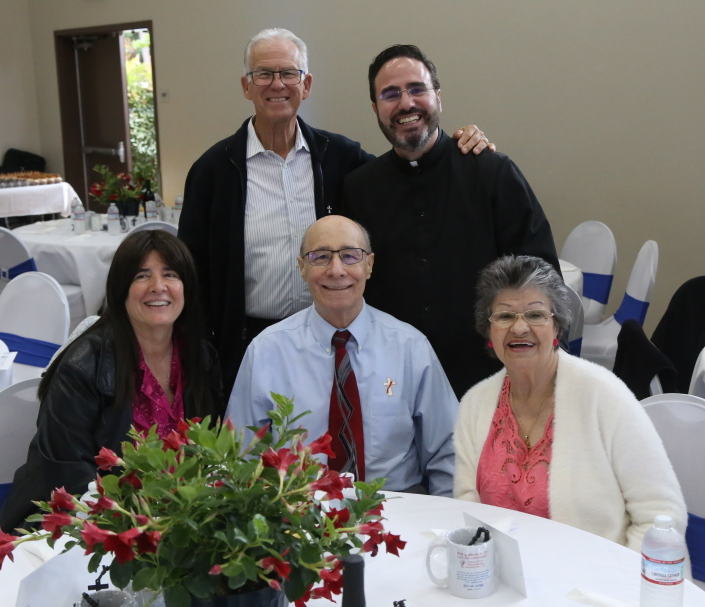Five people smiling at a table with flowers at a formal indoor gathering
