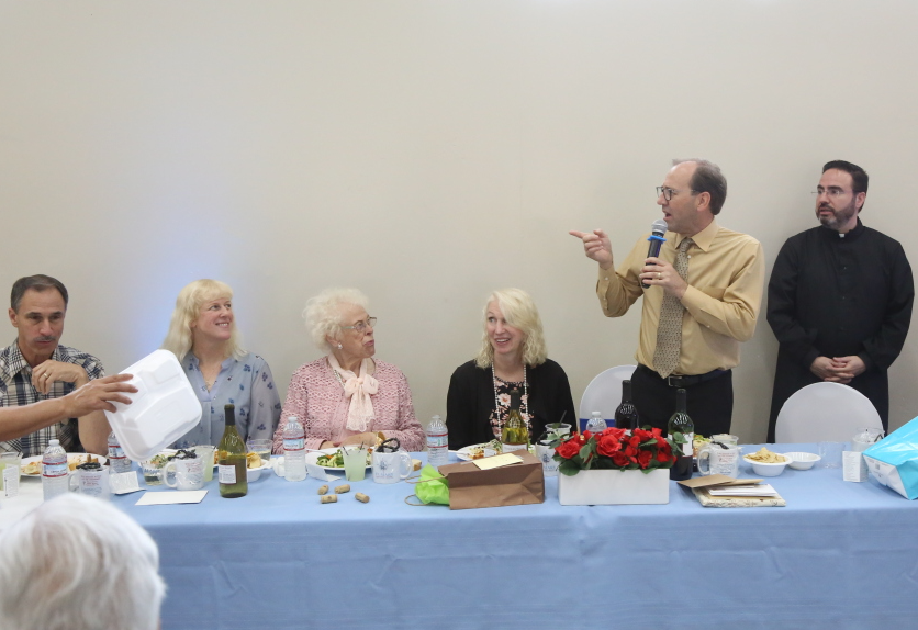 Man speaking at a banquet table while seated guests listen; blue tablecloth, food, flowers, and chairs