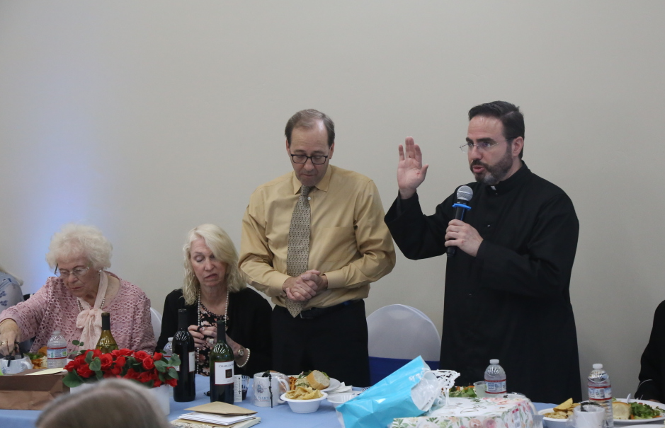 Man speaking into a microphone at a banquet table while others sit beside him, one person standing nearby.