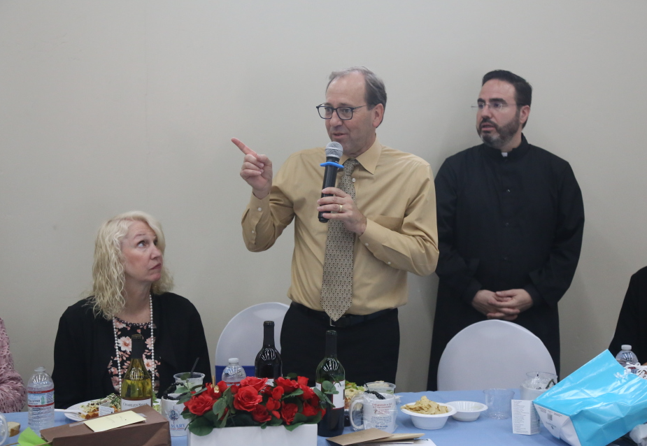 Man speaking into a microphone at a table, with two seated people and refreshments in a meeting room.