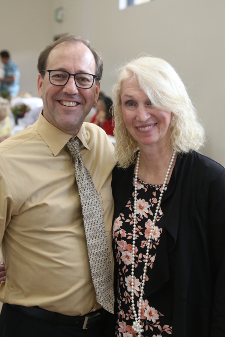 Two smiling people standing indoors, one in a tan shirt and patterned tie, the other in a black cardigan and floral top.