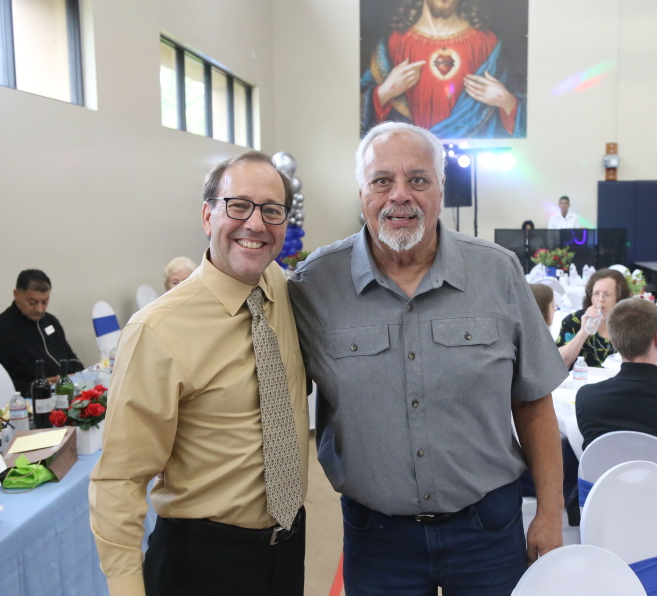 Two men smiling and posing together at an indoor event with tables and a religious painting in the background