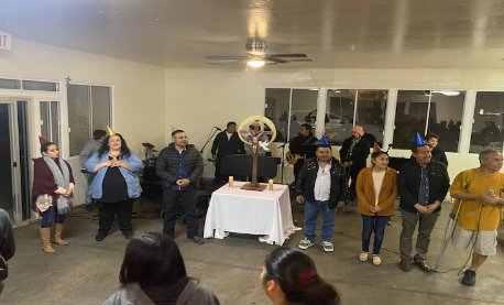 People standing in a hall, facing a decorated table and speaking at a community gathering