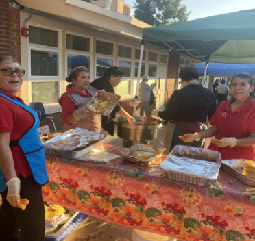 Volunteers serving food at an outdoor buffet table under a canopy near a building