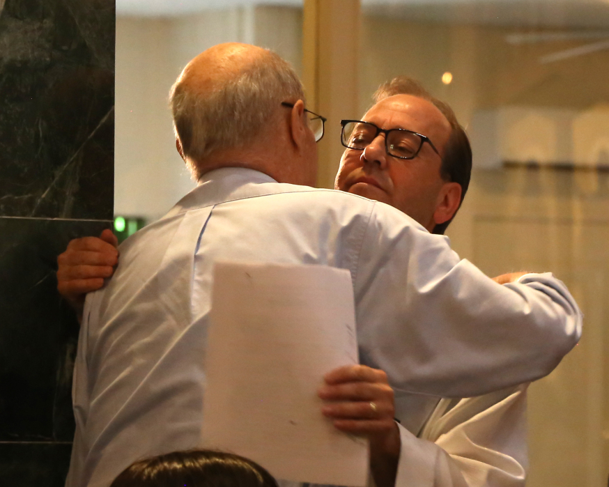 Two men in white shirts embrace in a hallway, one holding a sheet of paper behind the other’s back.