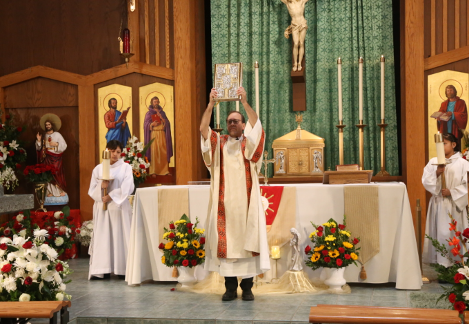 Priest elevates the Eucharist at altar during church mass, flanked by altar servers and floral arrangements.